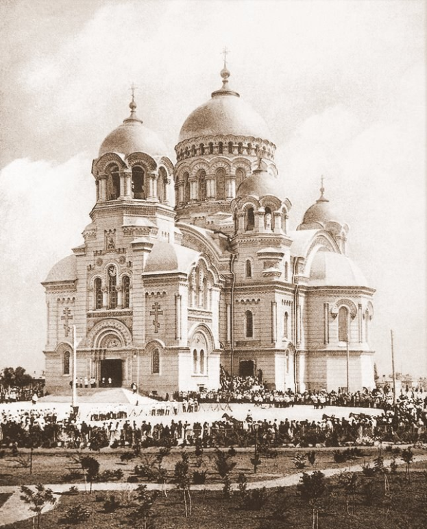 Schwarz-weiß-Fotografie der Erlöserkirche auf dem Blutsplatz in St. Petersburg, Russland, mit Menschen drumherum, Bäumen und Pfählen im Vordergrund und einem klaren Himmel im Hintergrund; Text ist unten sichtbar.