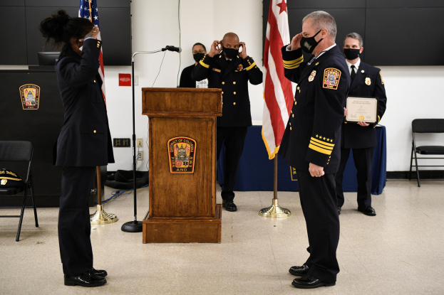 Gruppe uniformierter Personen in Masken vor einem Podium mit Mikrofon, umgeben von Flaggen und Möbeln, bei einer Vereidungszeremonie, bei der eine Person ein Zertifikat hält.