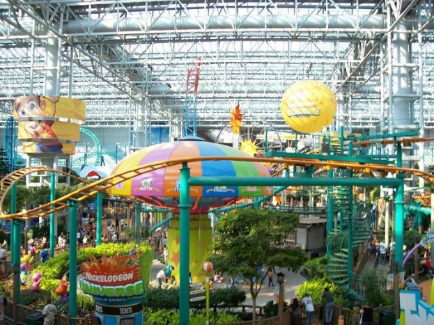 Ein großer Indoor-Wasserpark mit einer Achterbahn, die von Menschen, Bäumen, Pflanzen und einer farbenfrohen Beleuchtung umgeben ist.