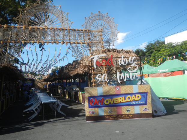 Eine KarnevalsSzene mit einem großen Riesenrad in der Mitte, umgeben von verschiedenen Buden, Sitzbereichen, dekorativen Elementen und Bäumen, mit einem bewölkten Himmel im Hintergrund.