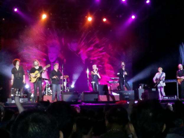 Eine Bühne mit Musikern, die Gitarren spielen und in Mikrofone singen, begleitet von Trommeln, unter hellen Scheinwerfern, mit einem Publikum im Vordergrund.