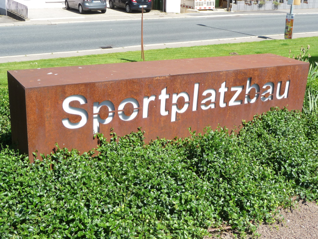 Ein Schild mit der Aufschrift "Sportplatzbau" steht vor einem Gebäude, umgeben von grünem Gras und Pflanzen, mit Fahrzeugen, Gebäuden, Pfählen und Brettern im Hintergrund.