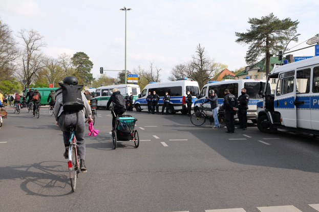 Eine Gruppe von Menschen, die auf Fahrr"aden eine Stra{\u00dfe entlangfahren, mit Polizeiwagen, anderen Fahrzeugen, B"aumen und Geb"auden im Hintergrund.