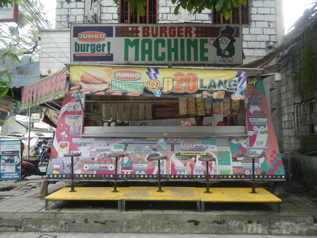 Ein leuchtend gelber und roter Foodtruck in Form einer Jumbo-Burger-Maschine, der auf einer von Gebäuden, Bäumen und Menschen umgebenen Straße geparkt ist.
