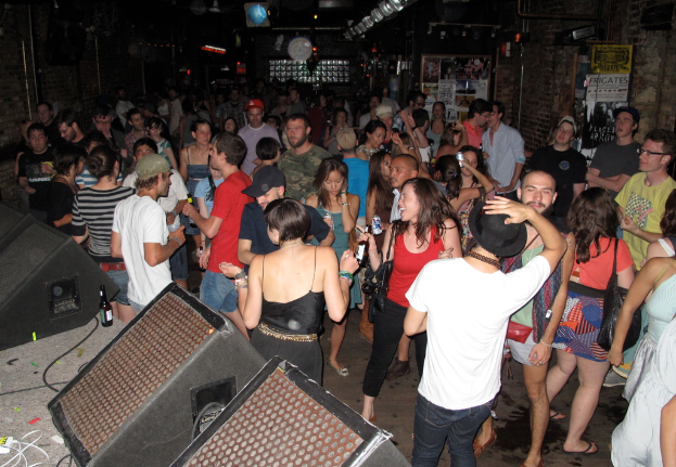 Eine Gruppe von Menschen, die tanzen und dabei einige Mützen tragen und Taschen halten, mit einem Lautsprecher, Kabeln, einer Flasche und Pfosten mit Text im Hintergrund.