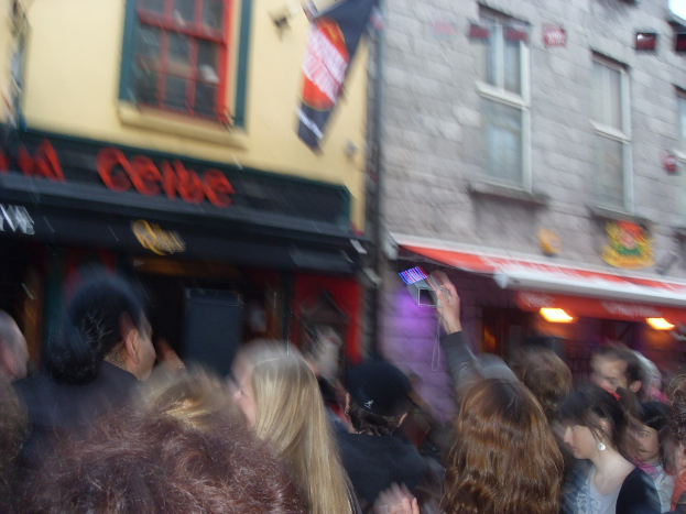 Ein unscharfer Bildausschnitt, der unten Menschen auf der Straße und oben Gebäude mit Lichtern, Fenstern und einer Flagge zeigt.