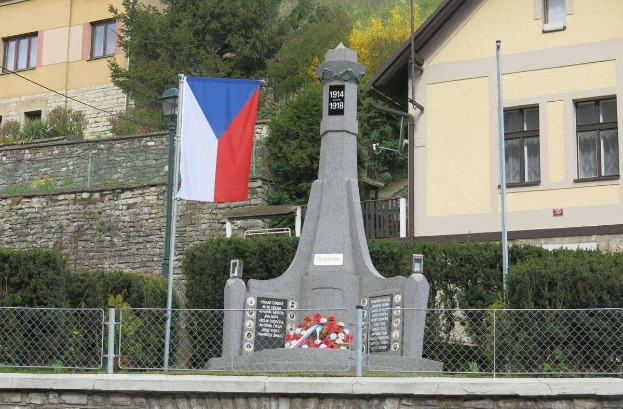 Ein Denkmal mit einer Fahne darauf, umgeben von einem Zaun, Pflanzen, Bäumen und einer Wand, dient als Gedenkstätte für die Opfer des Zweiten Weltkriegs mit eingravierter Inschrift.