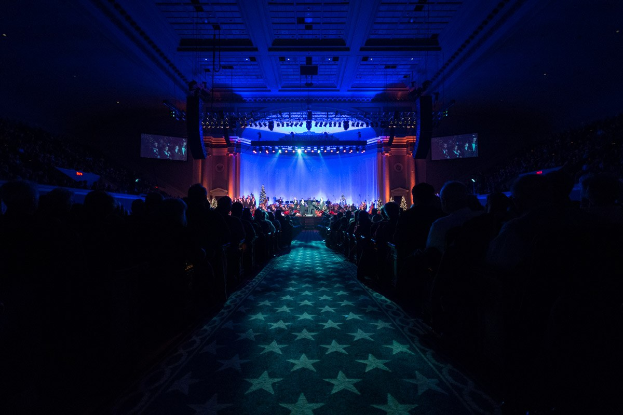 Ein großer Saal mit sitzenden Zuschauern, eine Bühne mit Musikern und Deckenleuchten und -fernseher, was auf ein Konzerthaus während der Nationalkonvention 2019 hindeutet.