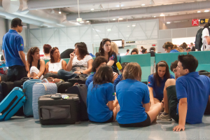 Ein belebter Flughafeninnenraum mit vielen wartenden Menschen, Gepäck auf der linken Seite und gläserne Türen mit Richtungsschildern im Hintergrund.
