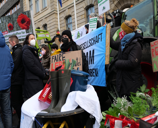Maskierte Menschen mit Protestschildern vor einem Lastwagen, mit einem Tisch mit Gegenständen, Topfpflanzen und einem beflaggten Gebäude im Hintergrund.