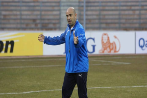 Ein Mann in einer blauen Jacke steht auf einem Fußballfeld, umgeben von Tafeln mit Text und Logos, entschlossen während eines Trainings.