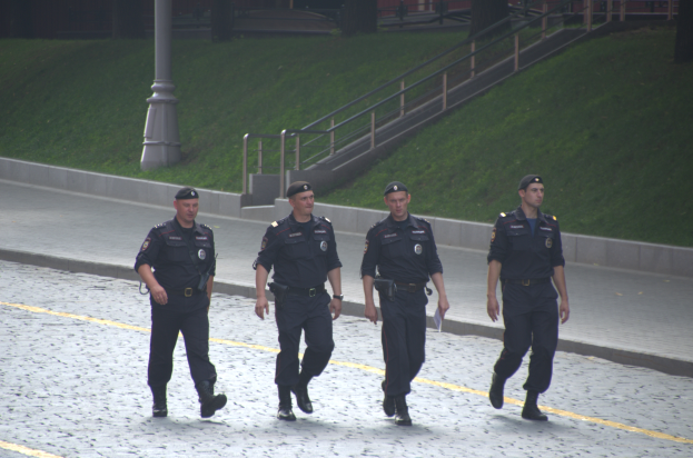 Vier Polizisten in Uniform gehen eine Straße entlang und halten Papiere in der Hand, mit einem Pfahl, Treppen, Geländern, Gras, Bäumen und einem Gebäude im Hintergrund.