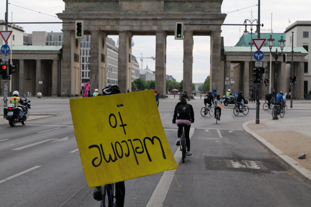 Eine Gruppe von Menschen in Helmen fährt mit Fahrrädern eine Straße entlang vor dem Brandenburger Tor in Berlin, Deutschland, mit einer Person, die eine gelbe Tafel hält, Laternenpfählen, Verkehrszeichen, Gebäuden, Bäumen und einem klaren blauen Himmel im Hintergrund.