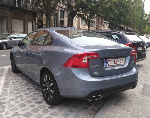 Ein Volvo S60 T5 AWD Inscription fährt auf einer Straße mit Gebäuden, Bäumen, Laternenmasten und einem klaren blauen Himmel.