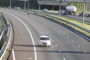 Ein Auto fährt auf einer Autobahn mit einem polizeilichen Fahrzeug am Rande, anderen Fahrzeugen, Geländern, Pfölen, Platten und Bäumen im Hintergrund.