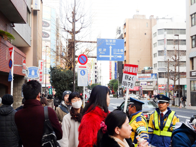 Eine belebte Straßenszene mit Menschen in verschiedenen Kleidungsstücken, darunter Mützen, eine Handtasche, eine Haarsträhne, ein Brett, ein Pfahl, Bäume, Gebäude mit Fenstern, eine Straße mit einer weißen Linie und einen weißen Himmel.