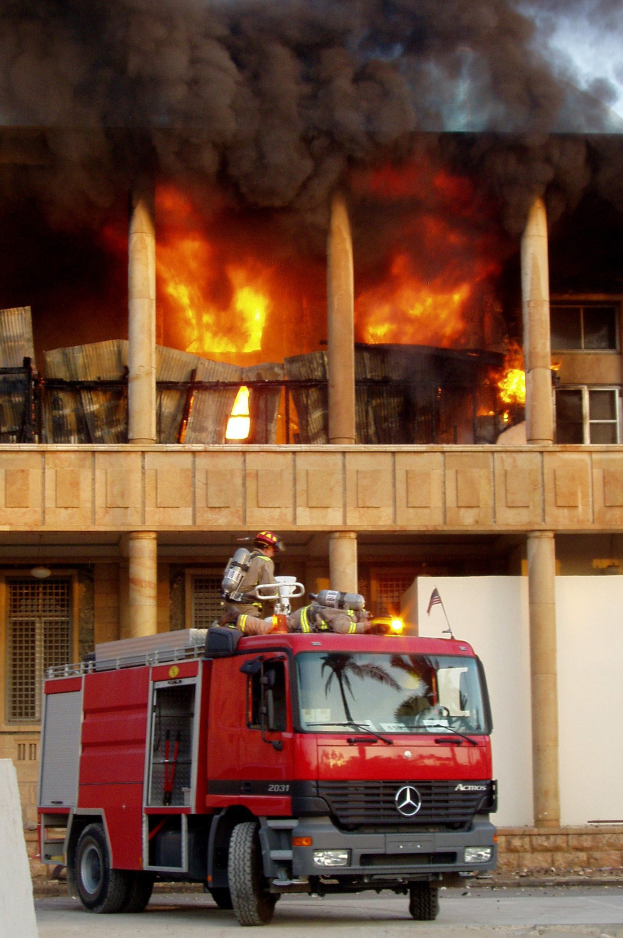 Feuerwehrl├Âschfahrzeug auf der Stra├če vor einem brennenden Geb├Ąude mit dickem Rauch, neben einer Wand.