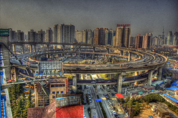 Eine Stadtansicht mit einer zentralen Ringstraße mit Fahrzeugen, einer unteren Straße mit Autos und Fahrrädern, zahlreichen Hochhäusern im Hintergrund, einem Himmel oben, Bäumen auf der linken unteren Seite und kleinen Häusern, die hier und da in der Stadt verteilt sind.