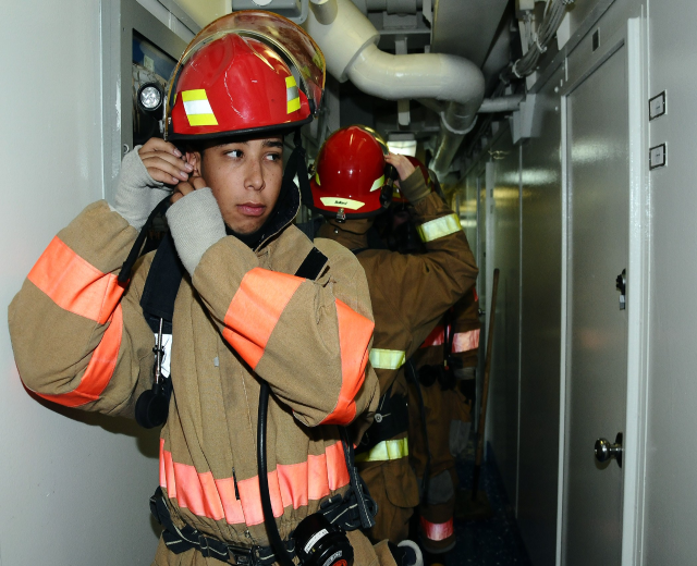Gruppe von Feuerwehrmännern in Uniform, die zusammen in einem Raum mit einer Tür auf der rechten Seite und sichtbaren Rohren im Hintergrund stehen.