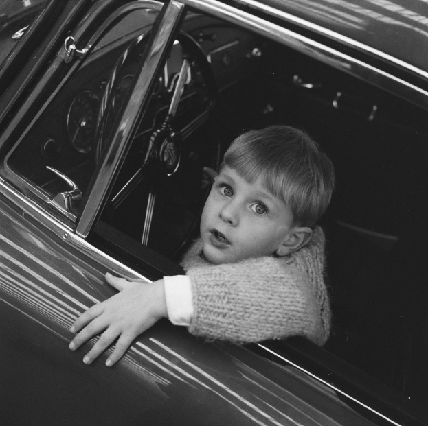Ein junger Junge in einem wei├čen Hemd und dunklen Hosen sitzt im Fahrersitz eines klassischen schwarzen Autos mit Chromakzenten und schaut aus dem Fenster mit einem neugierigen Ausdruck, H├Ąnde ruhen auf dem Lenkrad.
