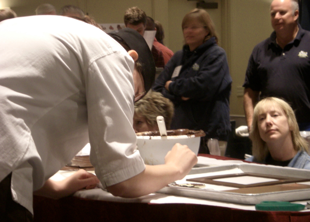 Eine Gruppe von Menschen vor einem Tisch mit einem Löffel, einer Schüssel und einem Tablett, mit einer Wand im Hintergrund, die eine Bibelszene zeigt.