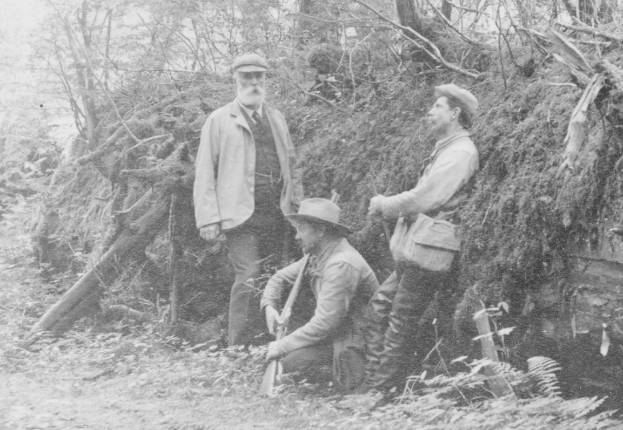 Drei Männer mit Mützen stehen zusammen in einem bewaldeten Gebiet, einer hält eine Waffe, auf einem Schwarz-Weiß-Bild.