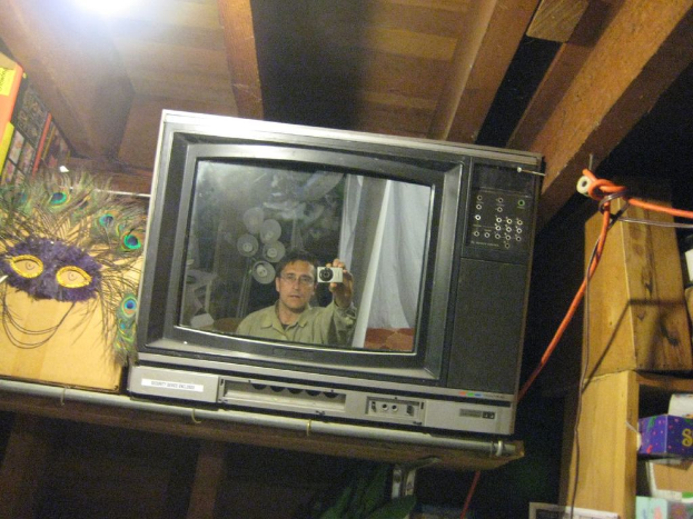 Ein Fernsehbildschirm mit einer Person umgeben von Pfauenfedern, BÃ¼chern und Kisten.