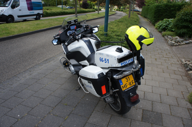 Ein auf der Seite der Straße geparktes Polizeimotorrad mit einem Helm darauf, umgeben von Bäumen, Pfählen, Gebäuden, Pflanzen, Gras, Steinen und anderen Fahrzeugen im Hintergrund.