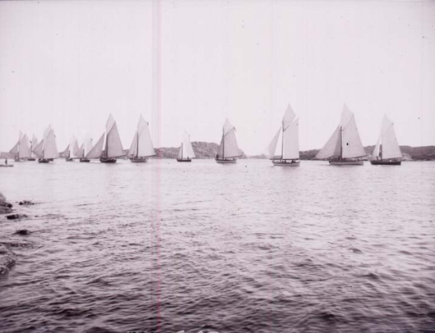 Schwarzes und weißes Foto von Segelbooten auf dem Wasser mit Hügeln im Hintergrund und einem klaren Himmel, beschriftet als ein Foto des New York Yacht Club.