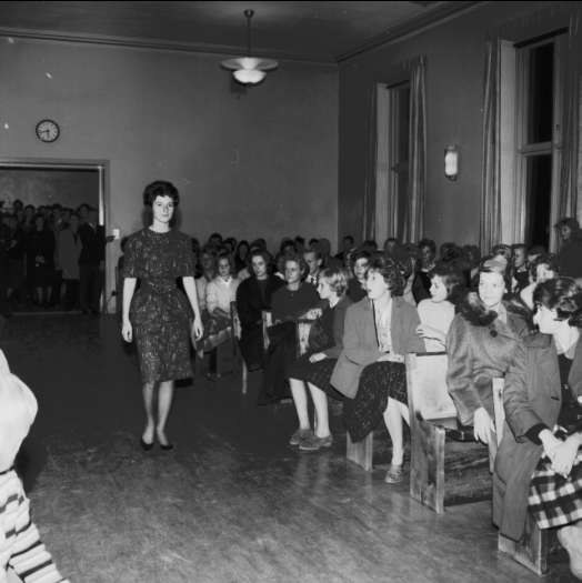 Eine Frau in einem schwarzen und weißen Kleid läuft über den Laufsteg, während das Publikum eine Modenschau zur Feier der Frauenwahlrecht beobachten.