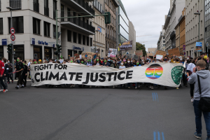 Eine Gruppe von Menschen marschiert mit einem Transparent, auf dem "Kampf für Klimagerechtigkeit" steht, eine Straße entlang, mit Gebäuden, Schildern, Verkehrsampeln und Bäumen an der Straße unter einem bewölkten Himmel.