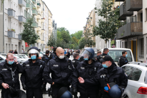 Polizisten in Masken und Helmen gehen eine Straße mit parkenden Autos gesäumt, flankiert von Gebäuden mit Balkonen und Geländern, unter einem klaren Himmel mit Bäumen im Hintergrund.