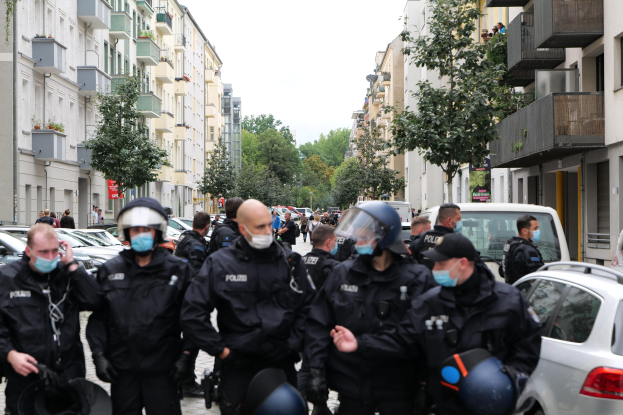 Polizisten in Masken und Helmen gehen eine Straße mit parkenden Autos gesäumt, flankiert von Gebäuden mit Balkonen und Geländern, unter einem klaren Himmel mit Bäumen im Hintergrund.