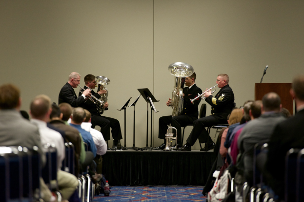 Eine Gruppe von Männern, die auf einer Bühne mit Notenpulten, einem Rednerpult und einem Mikrofon vor einem Publikum sitzen, das auf Stülhlen Platz genommen hat.
