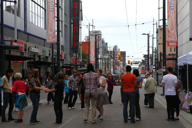Eine Gruppe von Menschen hält sich an den Händen auf einer Straße mit Gebäuden mit Geschäften im Erdgeschoss, Straßenlaternen und Masten mit Drähten und Werbetafeln, mit Zelten auf dem rechten Gehweg.