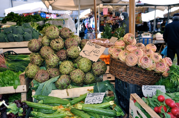 Körbe gefüllt mit Obst, Gemüse und kleinen Tafeln mit Text, mit Zelten und ein paar Menschen im Hintergrund.