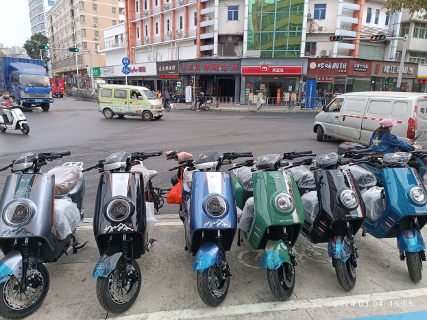 Eine Reihe von Elektro-Scootern, die auf einer Straße geparkt sind, mit einer Person in einer Mütze davor, vor einer Kulisse von Gebäuden, Ampeln, Bäumen und einem klaren blauen Himmel.