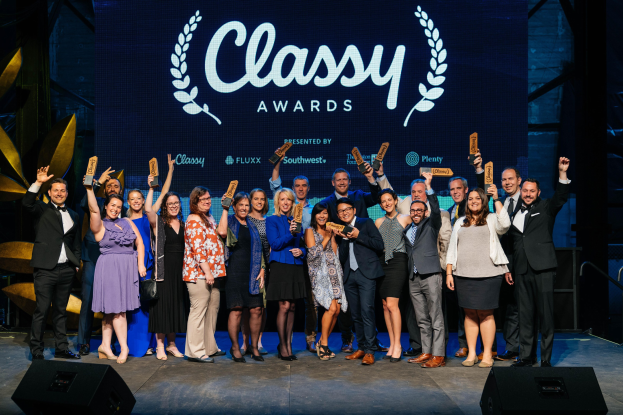 Gruppe von Menschen auf einer Bühne mit Auszeichnungen, mit Lautsprechern darunter und einem Bildschirm im Hintergrund, der 'Classy Awards 2019' anzeigt.