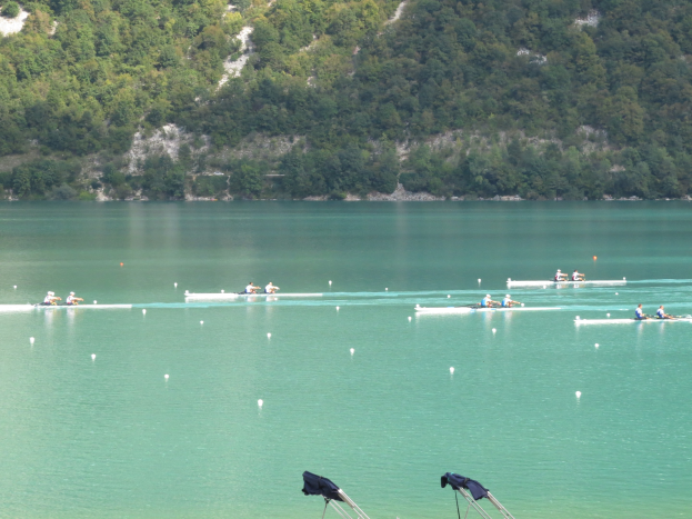 Eine Gruppe von Menschen, die bei einem Wettbewerb auf einem Gewässer rudern, mit Bäumen und einem Hügel im Hintergrund und Kleidung, die über Stühle am Wasserrand gehängt ist.