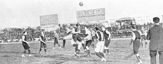 Ein Schwarz-Wei├č-Bild von einer Gruppe von M├Ąnnern, die auf einem Feld F├╝├čball spielen, mit einem Ball in der Luft und Zuschauern im Hintergrund, mit Schildern, die Text ├╝ber das Spiel anzeigen.