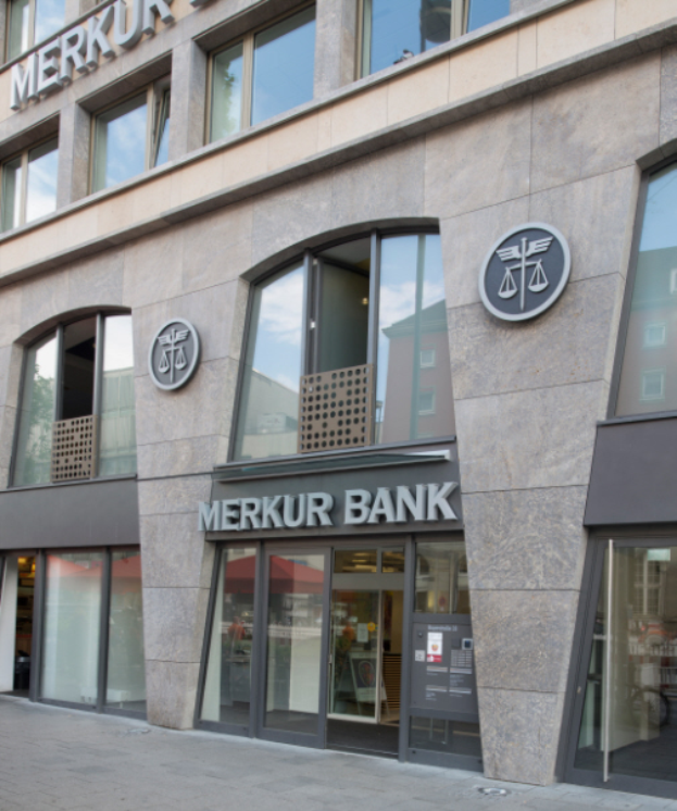 Ein modernes Gebäude mit Glaswänden und -türen, auf dem die Aufschrift "Merkur Bank" zu sehen ist, das neben einer Straße steht.