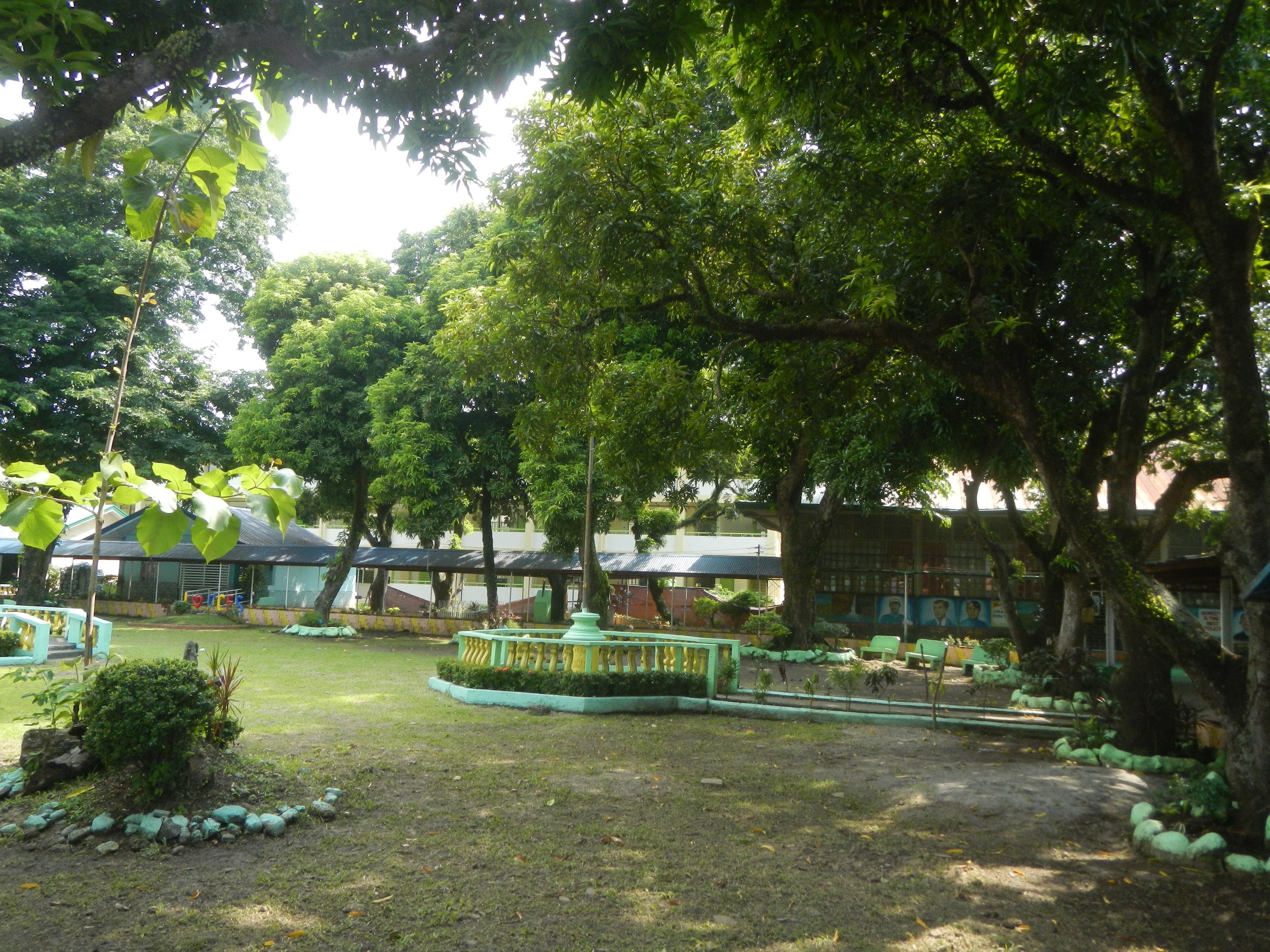 Ein Park mit grünem Gras, Pflanzen, Bäumen, einem zentralen Brunnen, Gebäuden im Hintergrund und einem klaren blauen Himmel.