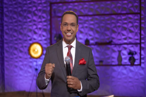 Stephen Colbert, der einen Anzug, Krawatte und Brille trägt, steht auf einer Bühne und hält ein Mikrofon in der Hand, während im Hintergrund eine Wand und verschiedene Gegenstände zu sehen sind.