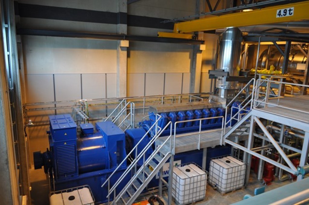 Eine Fabrikinnensicht mit Trägern, einer Treppe, Geländer, Containern, Lichtern, Röhren und Wänden.