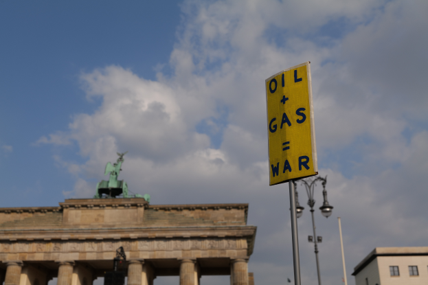 Das Brandenburgertor in Berlin, Deutschland, mit einem gelben Schild, das "Öl- und Gaskrieg" lesenbar ist, im Vordergrund, Gebäuden, Polen, Lichtern, einer Statue und einem bewölkten Himmel im Hintergrund.