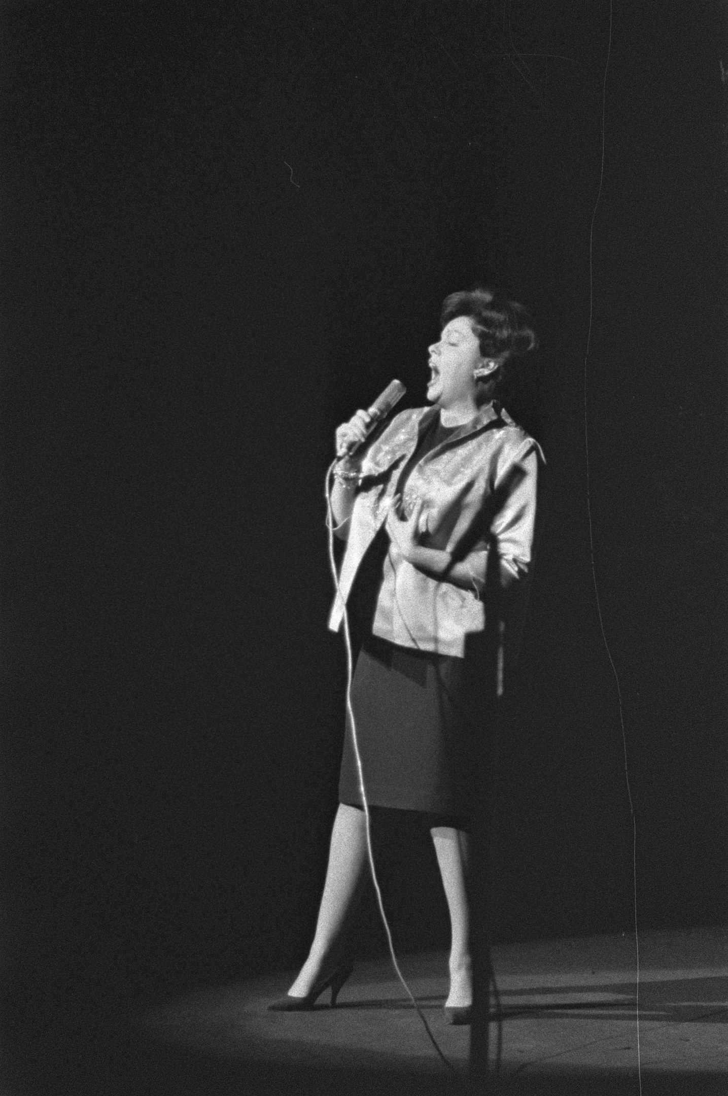 Eine Frau in einem schwarz-weißen Outfit aus den 1950er-Jahren singt fröhlich auf einer von einem Scheinwerfer beleuchteten Bühne in ein Mikrofon, ihre Haare sind im klassischen 50er-Jahre-Stil frisiert.