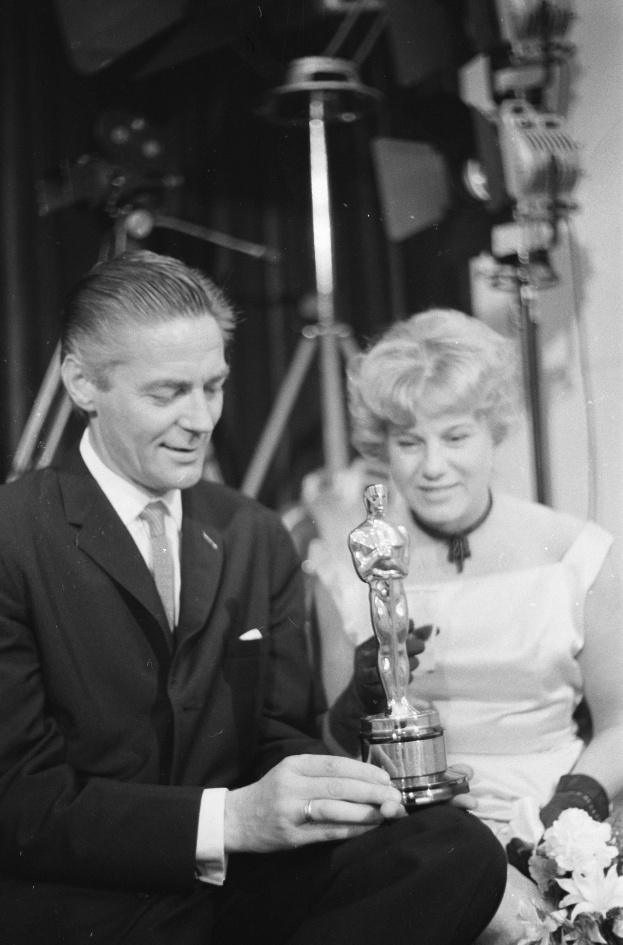 Ein Mann in einem schwarzen Blazer, weißem Hemd und Krawatte hält eine Oscar-Statue in der Hand und sitzt neben einer Frau, die einen Blumenstrauß hält, während Schwarz-Weiß-Lichter die Szene erhellen.