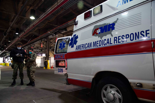Ein Rettungswagen der American Medical Response (AMR) steht in einem Gebäude, vor ihm stehen zwei maskierte Personen, links ein Fahrzeug und im Hintergrund mehrere Personen, Lichter und Rohre.