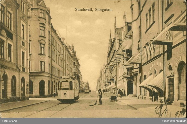 Ein altes Foto einer Stadtstraße mit einer Tram, Gebäuden, Passanten, Fahrrädern und einem bewölkten Himmel, mit Text oben und unten.