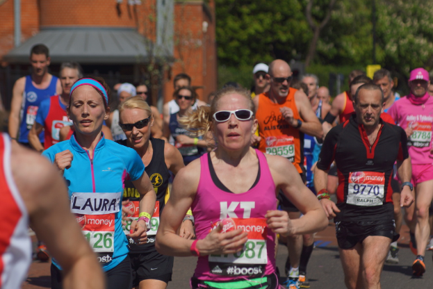 Mehrere Menschen joggen in bunten Shirts, mit einem Gebäude links und einem Baum rechts.